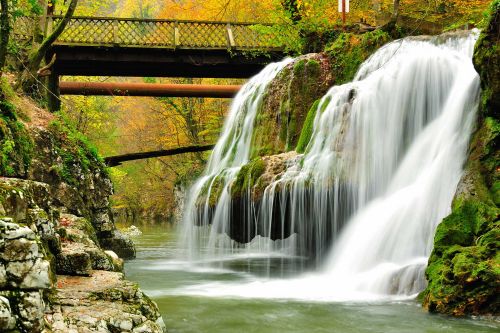 A smaller waterfall flows into a river in a forest area. A small footbridge and a pipeline cross the river.